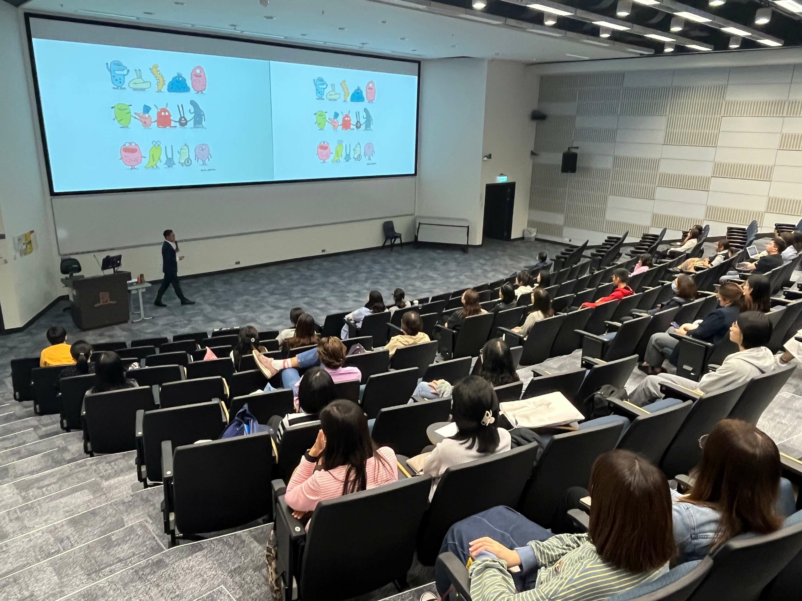 Participants in the lecture hall listening to the guest and looking at the screen with illustration of colourful characters.