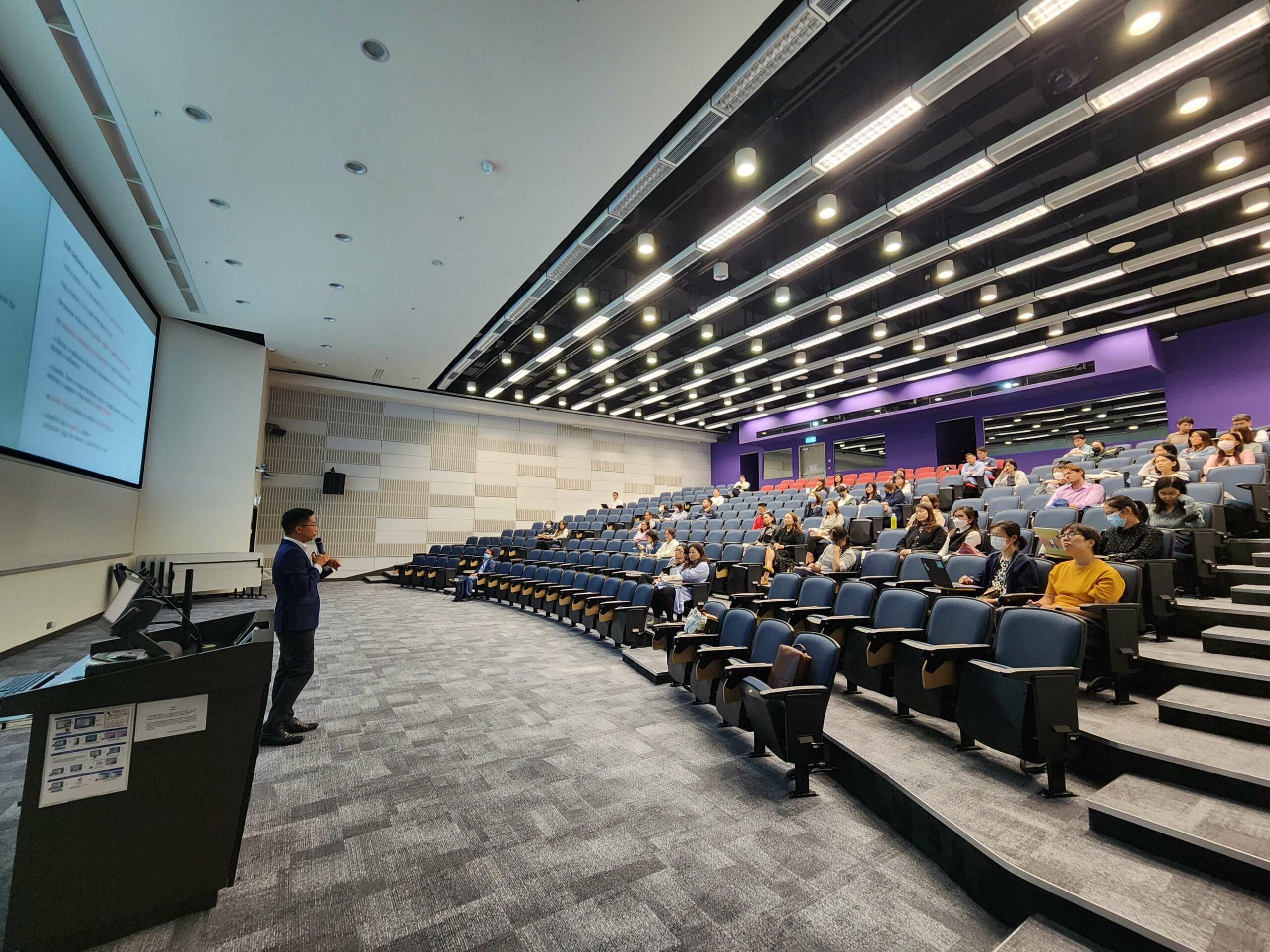 The guest speaker stands in the middle of the stage while participants listen and take notes on their devices.